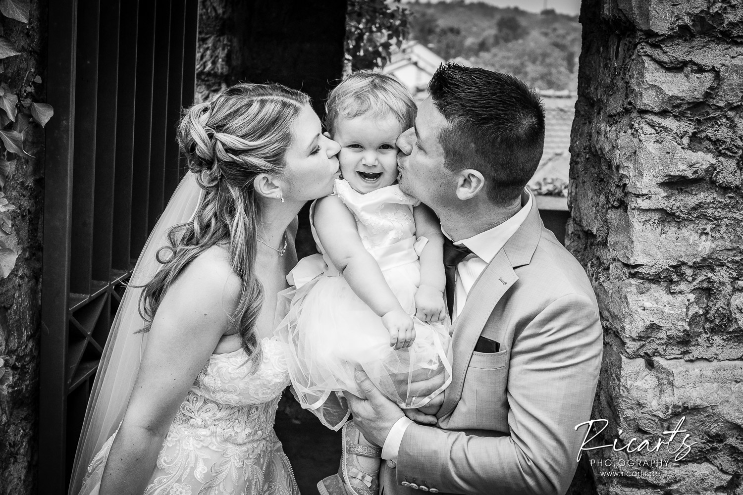 Hochzeitspaar-mit-kleinem-Kind-auf-Arm-beide-küssen-das-kind
