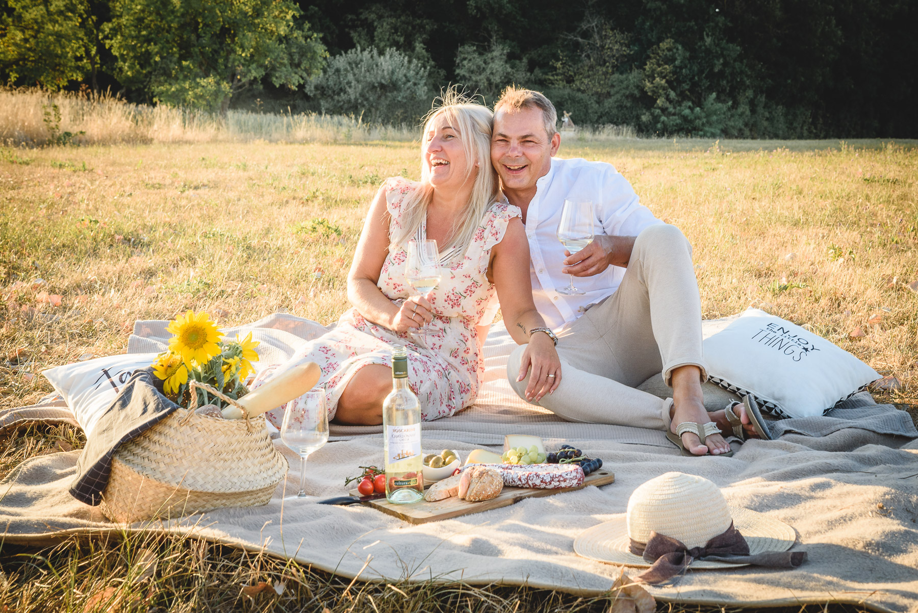 Paar sitzt lachend auf einer Picknickdecke mit einem Weinglas in der Hand auf einer Sommerwiese